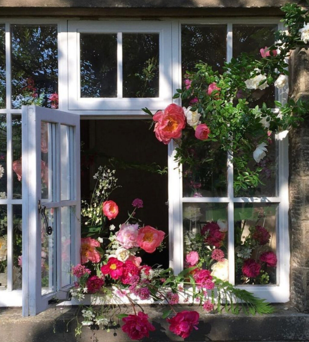 Roses clamber around the window on a stone built house, and an arrangement made with peonies and red valerian cascades through the open window. Photo: Beamsley Blooms.