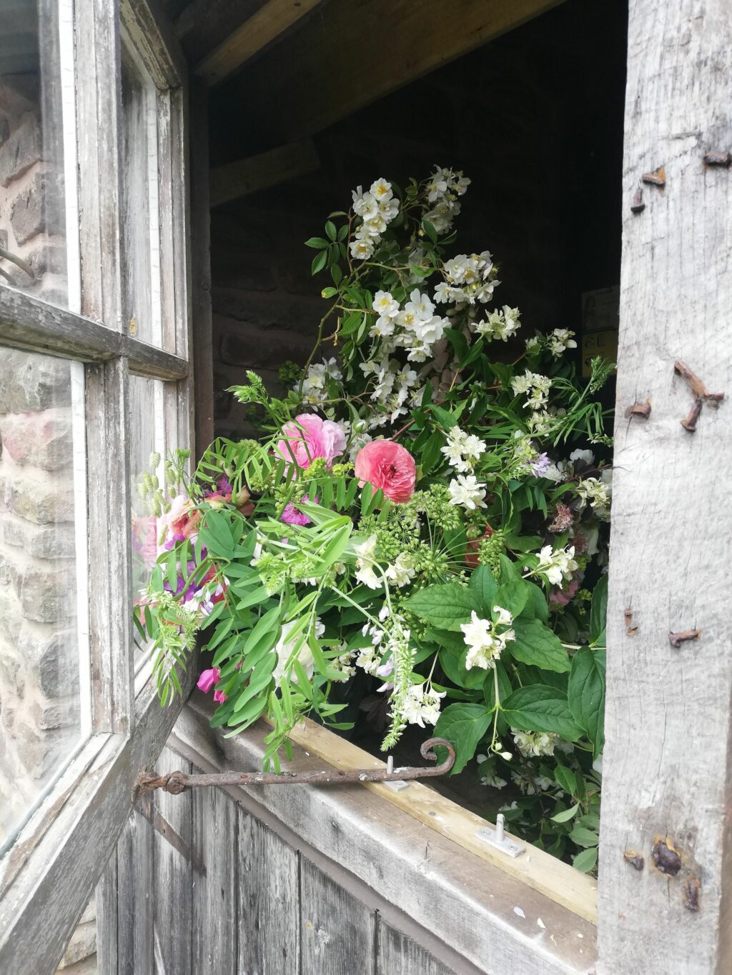 A late spring flower arrangement with bursting green foliage and freshly cut ranunculus perches on a wooden stable door at Farhill Flowers' flower farm in Wales.