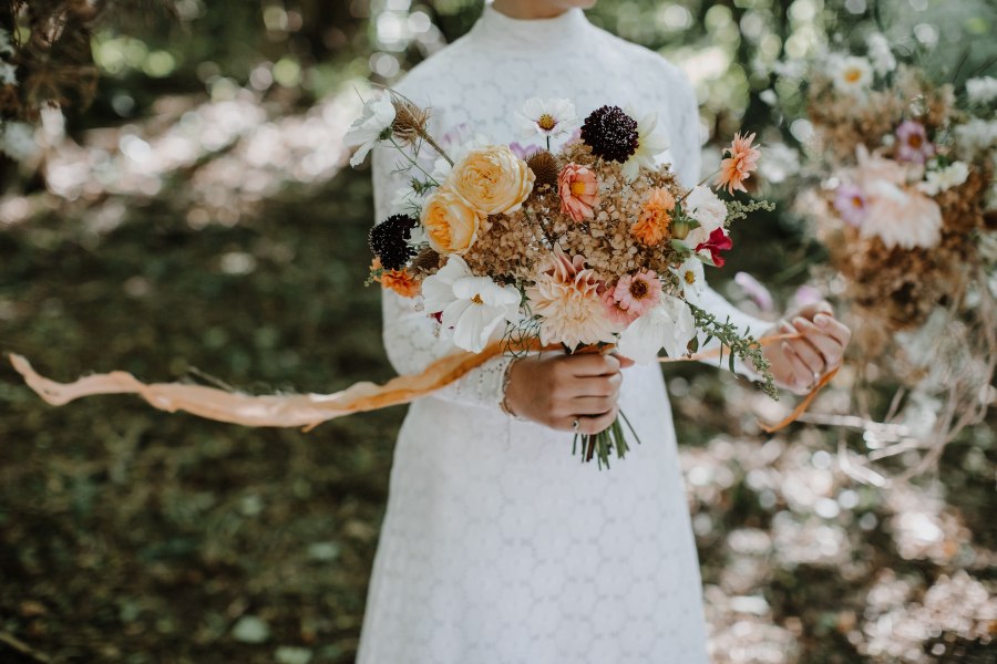 A romantic autumn bouquet with dahlias and dried hydrangeas with floaty silk ribbon held by a bride. Botanical Yard.