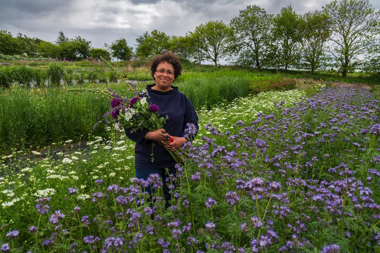 Cel Roberston of Forever Green Flower Company stands in her late spring flower field holding a bouquet of purple alliums and pink foxgloves. The beds in front of her are awash with blue phacaelia and white love in a mist.