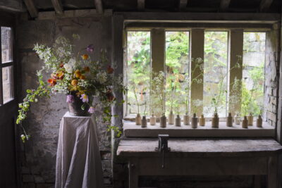 Simply By Arrangement's delicate window display consist of a parade of local British flowers in tiny vessels in mulllioned window, with a large tumbling arrangement on a linen covered plinth.