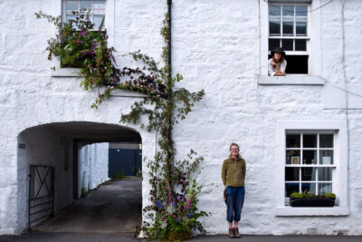 An installation of flowers climbs up the exterior wall at Tomnah'a Flowers, Scotland whilst the florists who created it lean out of windows and against the wall.