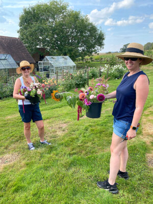 Visitors to the Ledbury Flower Farmer hold buckets of bright dahlias and cascading amaranths in the sunny flower field.