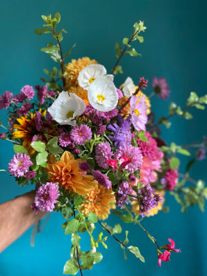 A joyous bouquet of white cosmos, pink cosmos, bright orange dahlias and foliage pops with colour against a turquoise background. 